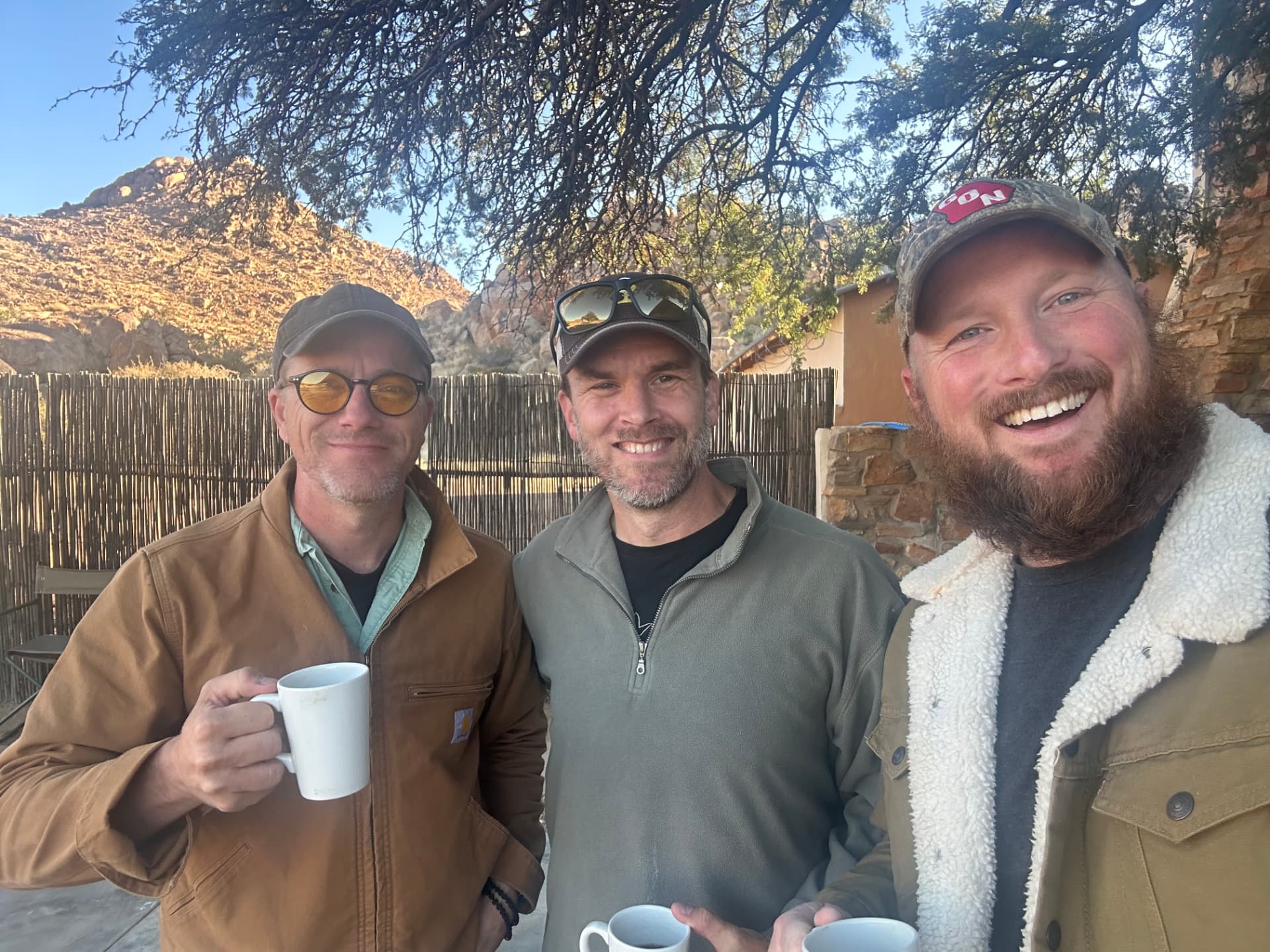 Three men laughing over morning coffee in a rugged mountain camp
