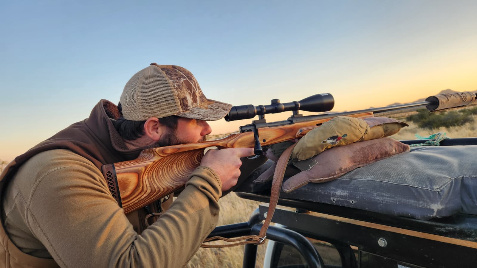 A hunter steadying a rifle on shooting bags across a vehicle rail at first light