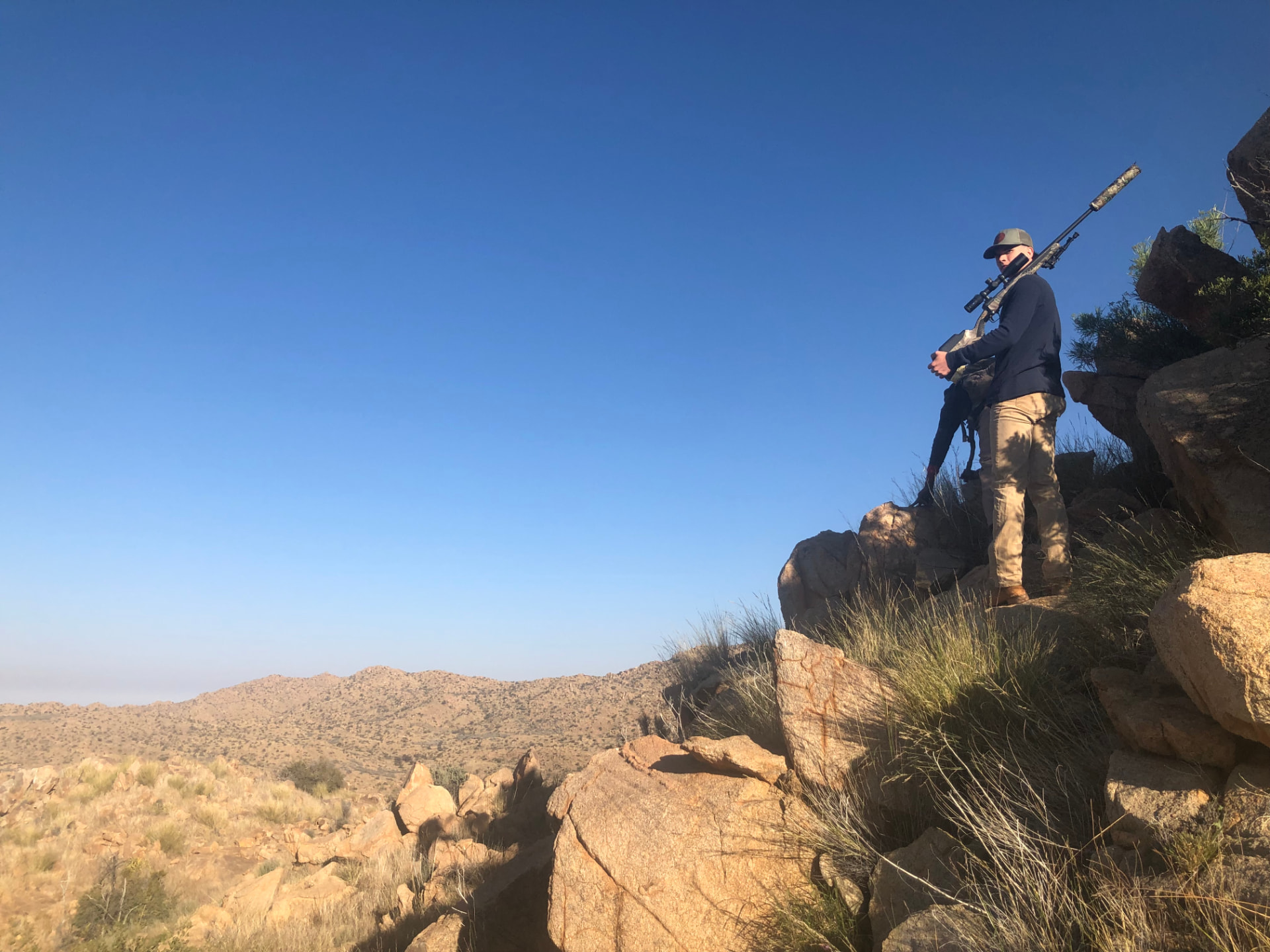 A hunter standing on a rocky outcrop overlooking a vast arid valley, rifle in hand