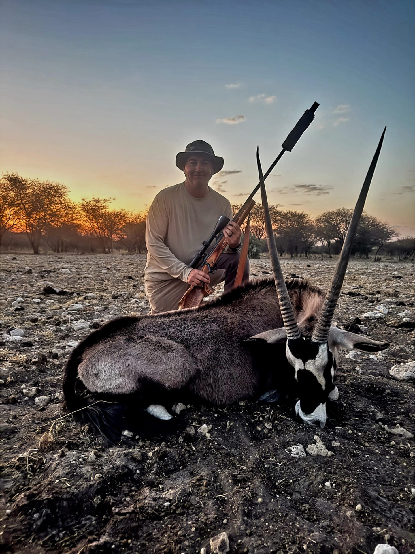 A hunter kneeling beside a harvested oryx in a rocky field at sunset