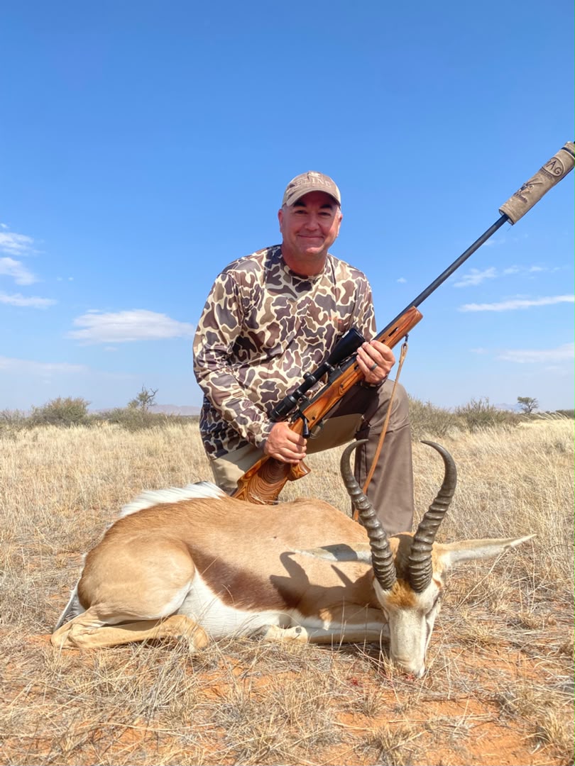 A hunter kneeling in dry brush with a harvested springbok and rifle