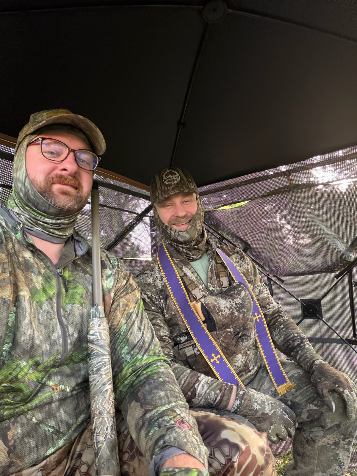 A priest in camouflage wearing a purple confessional stole hearing confession beside a hunter inside a ground blind